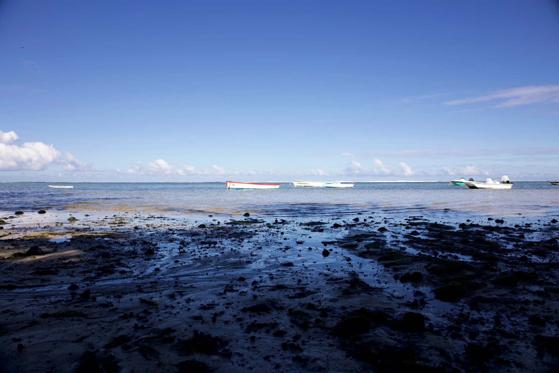 Small boats anchored in calm shallow water under a wide blue sky with low clouds, on a rocky tidal flat at Rivière Noire, Mauritius