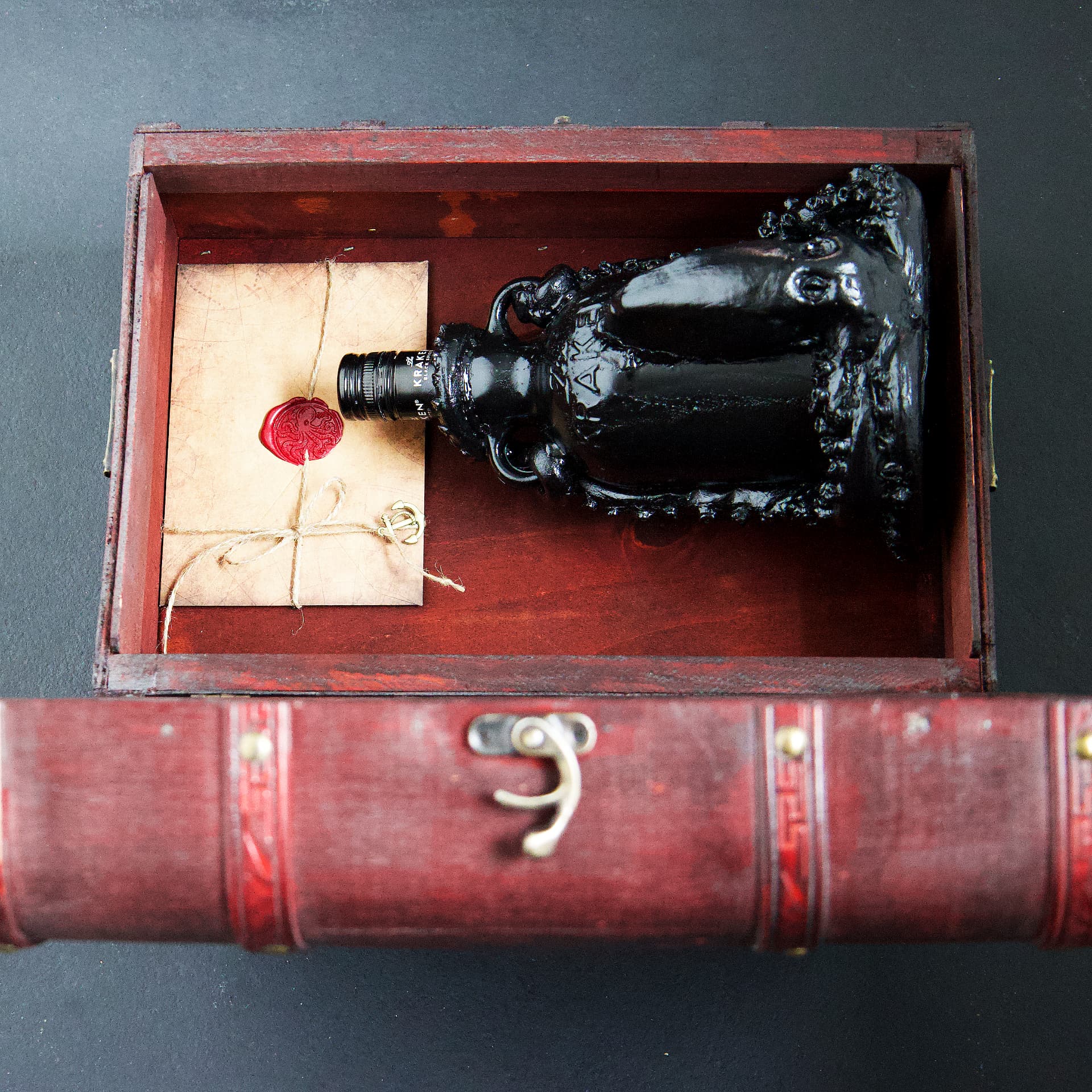 A black Kraken rum bottle sculpted with dark tentacles, lying inside an open vintage wooden chest beside a wax-sealed letter.