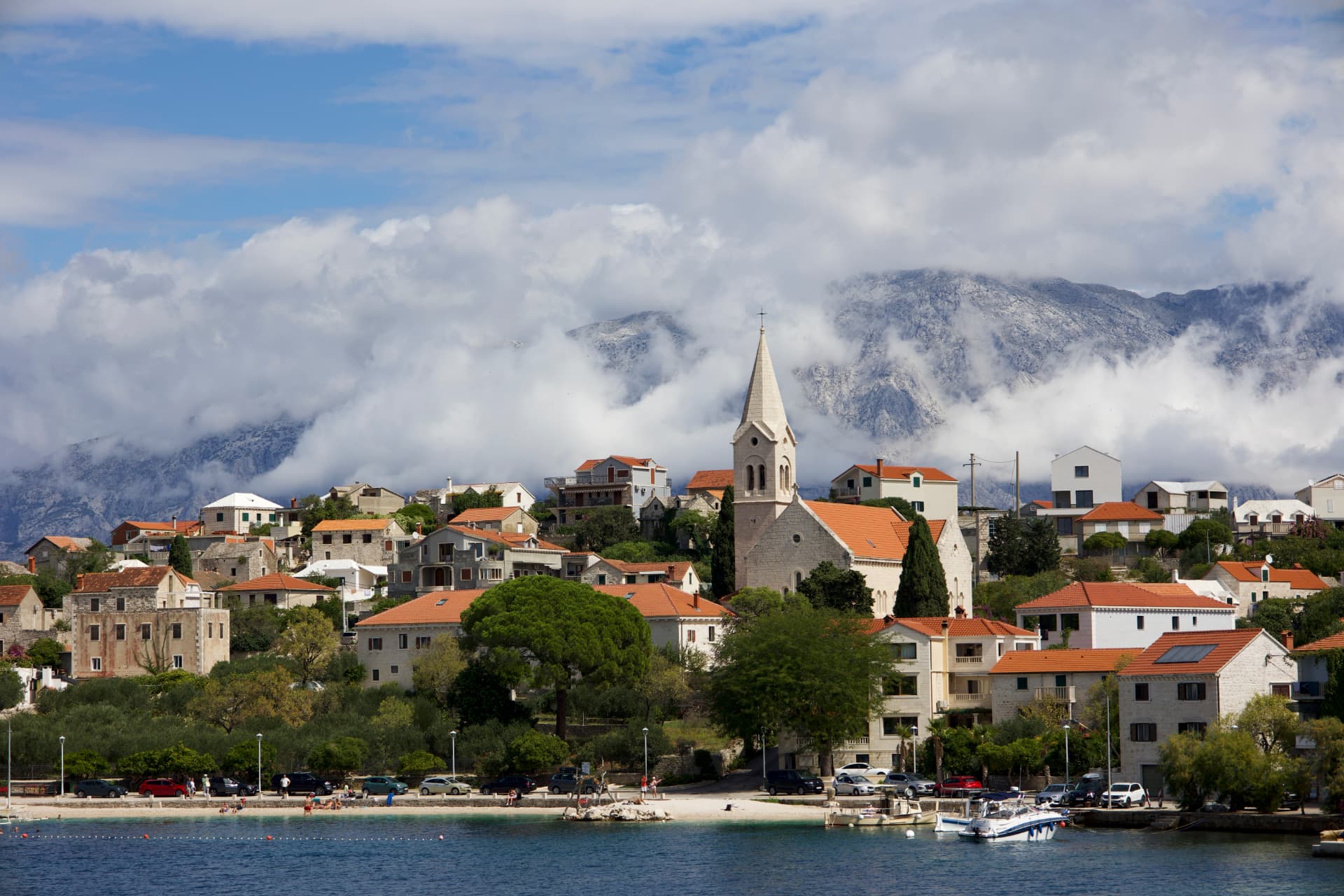 A coastal village with a stone church and red-tiled roofs on Brač Island, with mountains and clouds across the water, Croatia