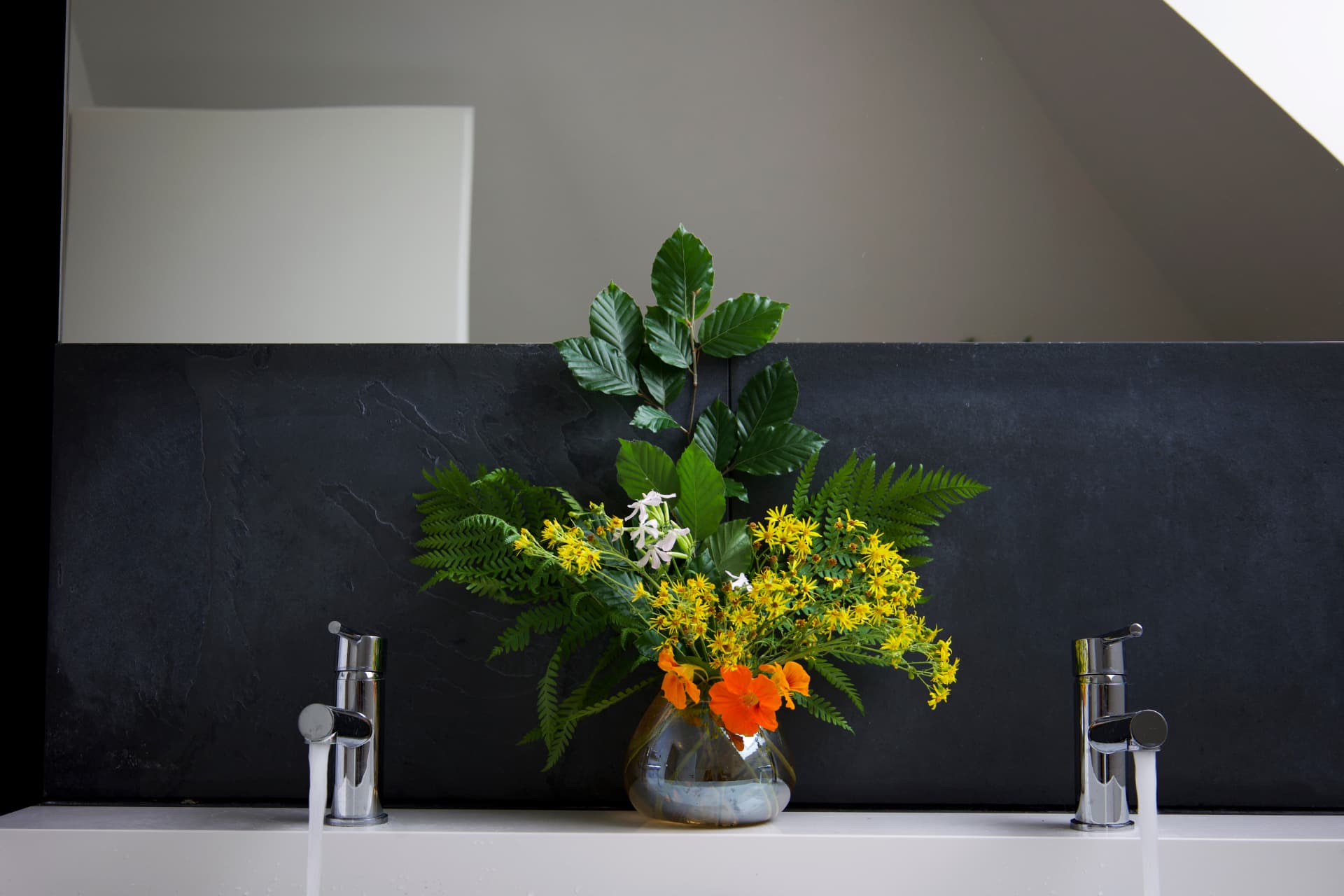 A glass vase with yellow and orange flowers, ferns, and green beech branches against slate.