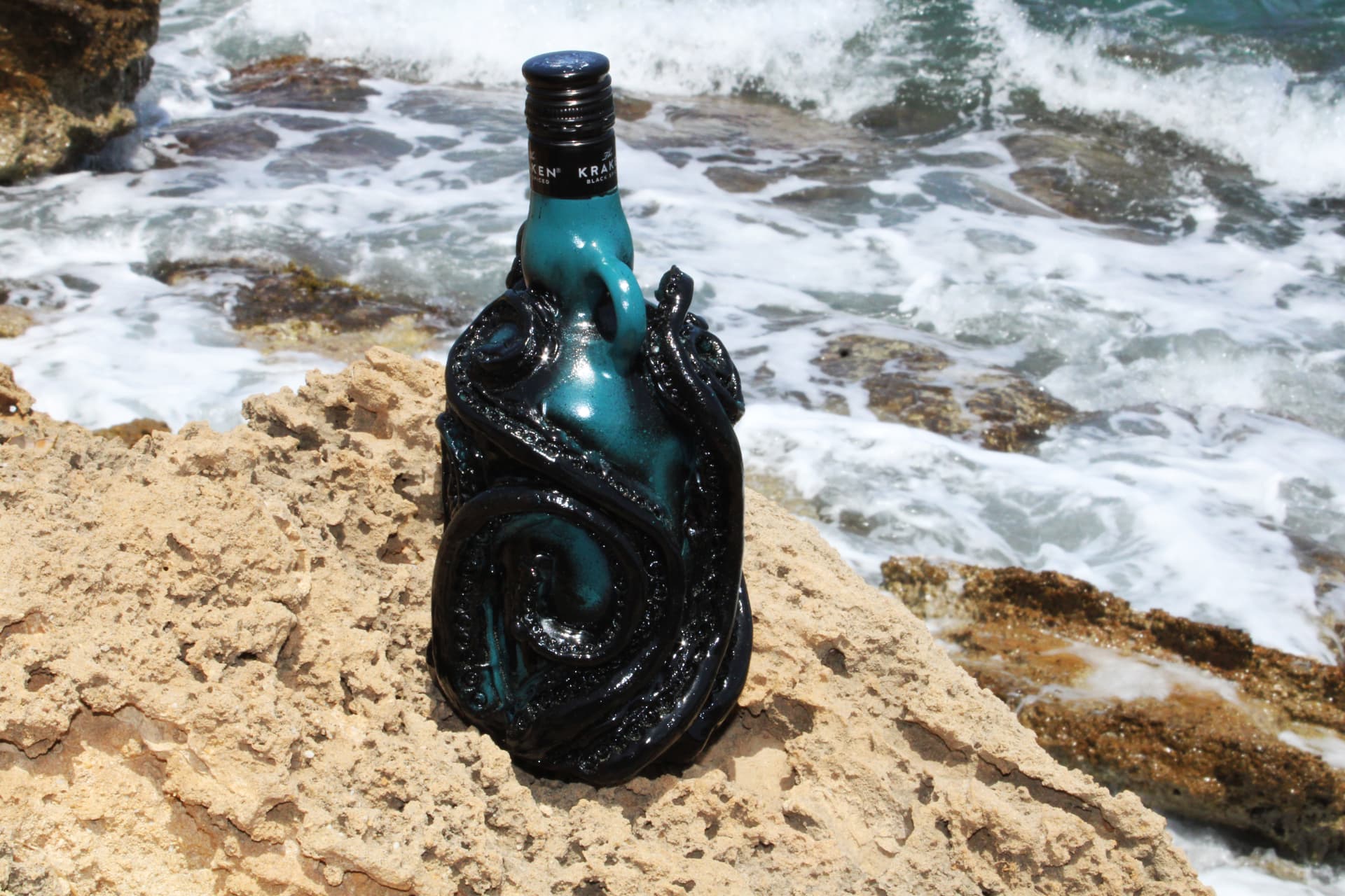 A teal Kraken rum bottle sculpted with black tentacles, standing on a rocky shore with waves behind it.