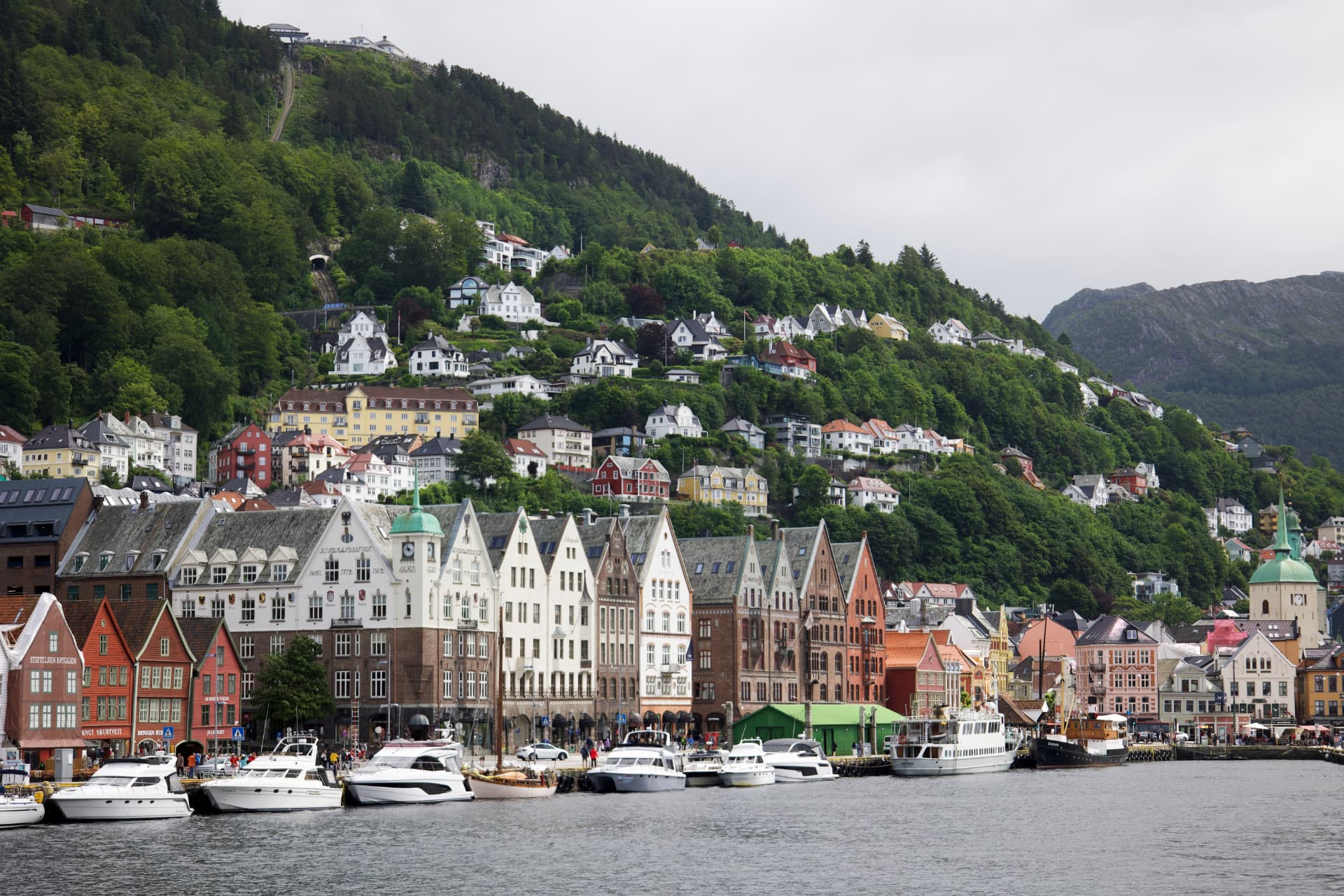 Colourful historic warehouses and boats along Bergen harbour with green hills and Mount Fløyen rising behind, Norway