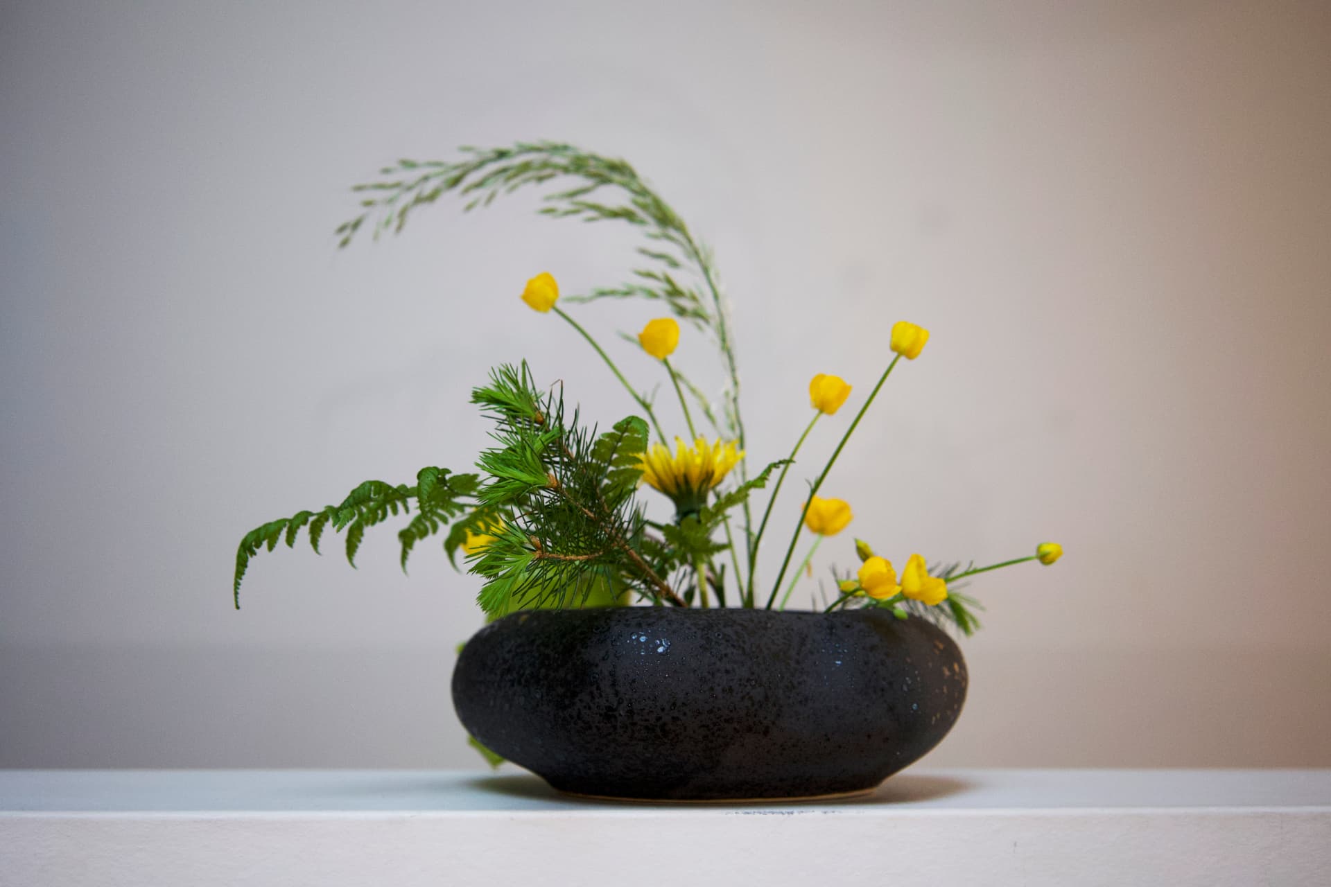 A low black ceramic bowl with yellow buttercups, ferns, and pine branches.