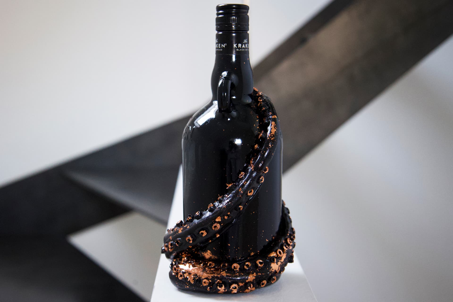 A black Kraken rum bottle decorated with sculpted black tentacles and copper-gilded details, standing on a white surface against a soft grey background.