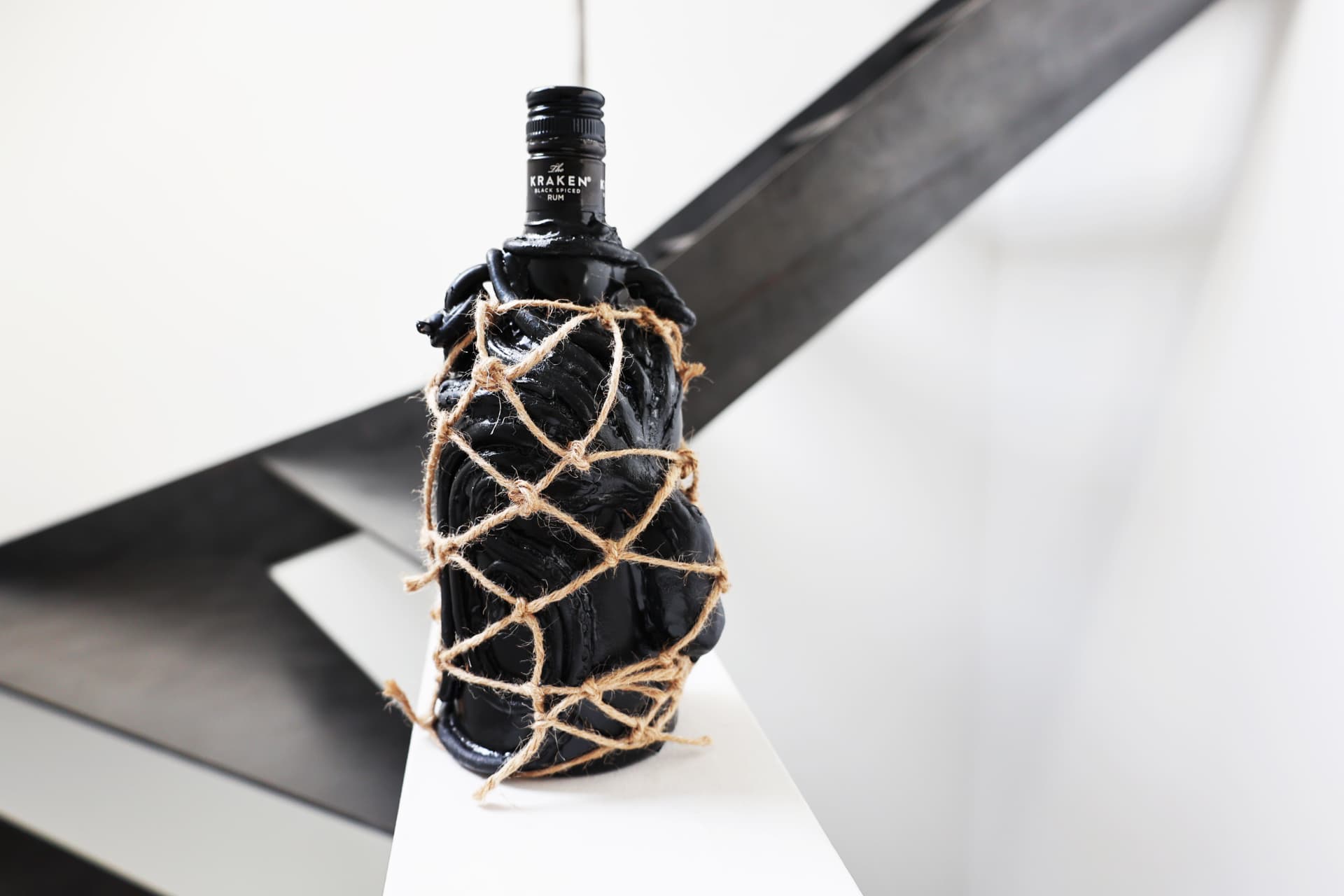 A black Kraken rum bottle wrapped with sculpted tentacles and twine netting, displayed on a white surface against a geometric black-and-white background.
