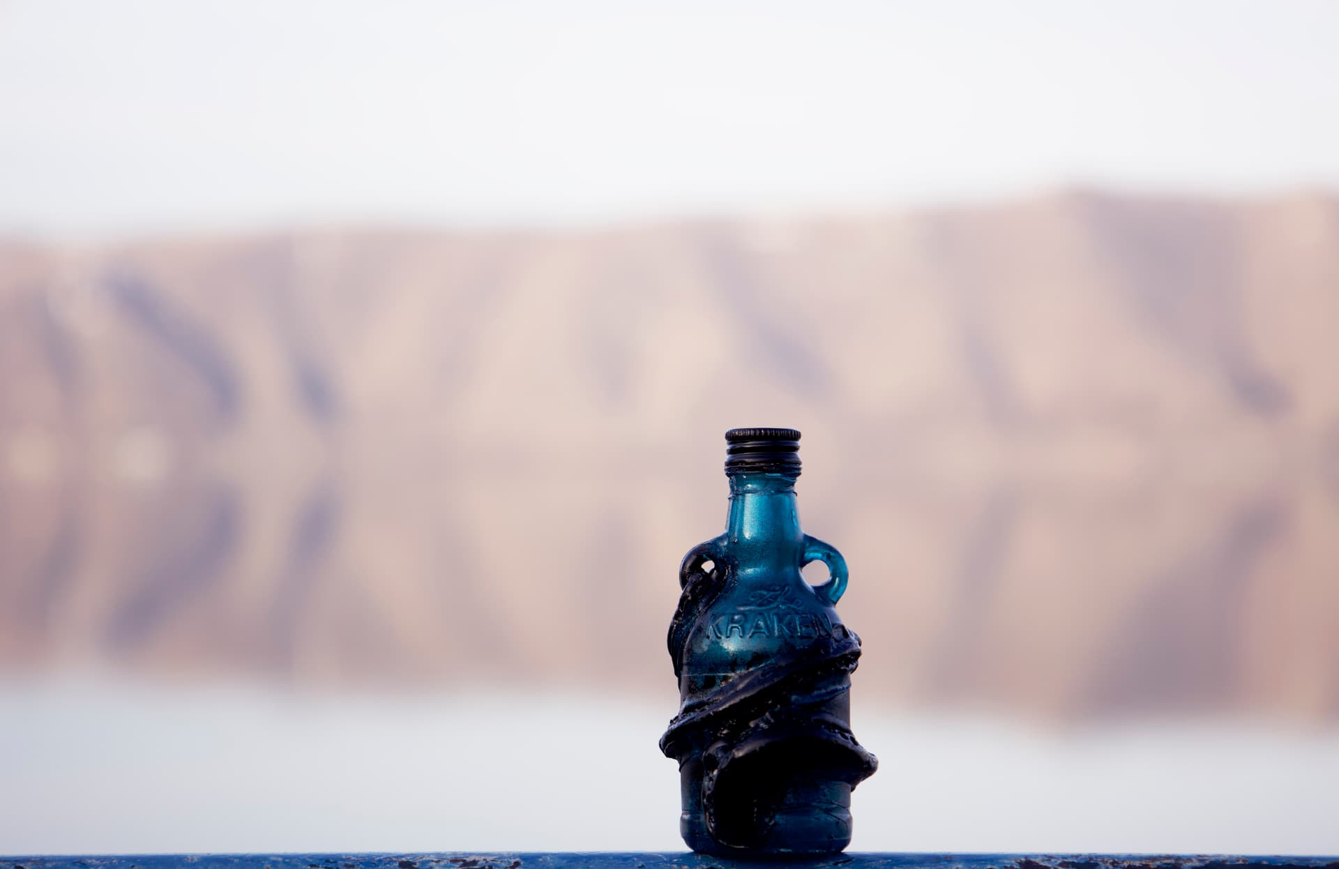 A small blue Kraken rum bottle wrapped with sculpted black tentacles, standing on a blue railing with a soft rocky shoreline behind it.