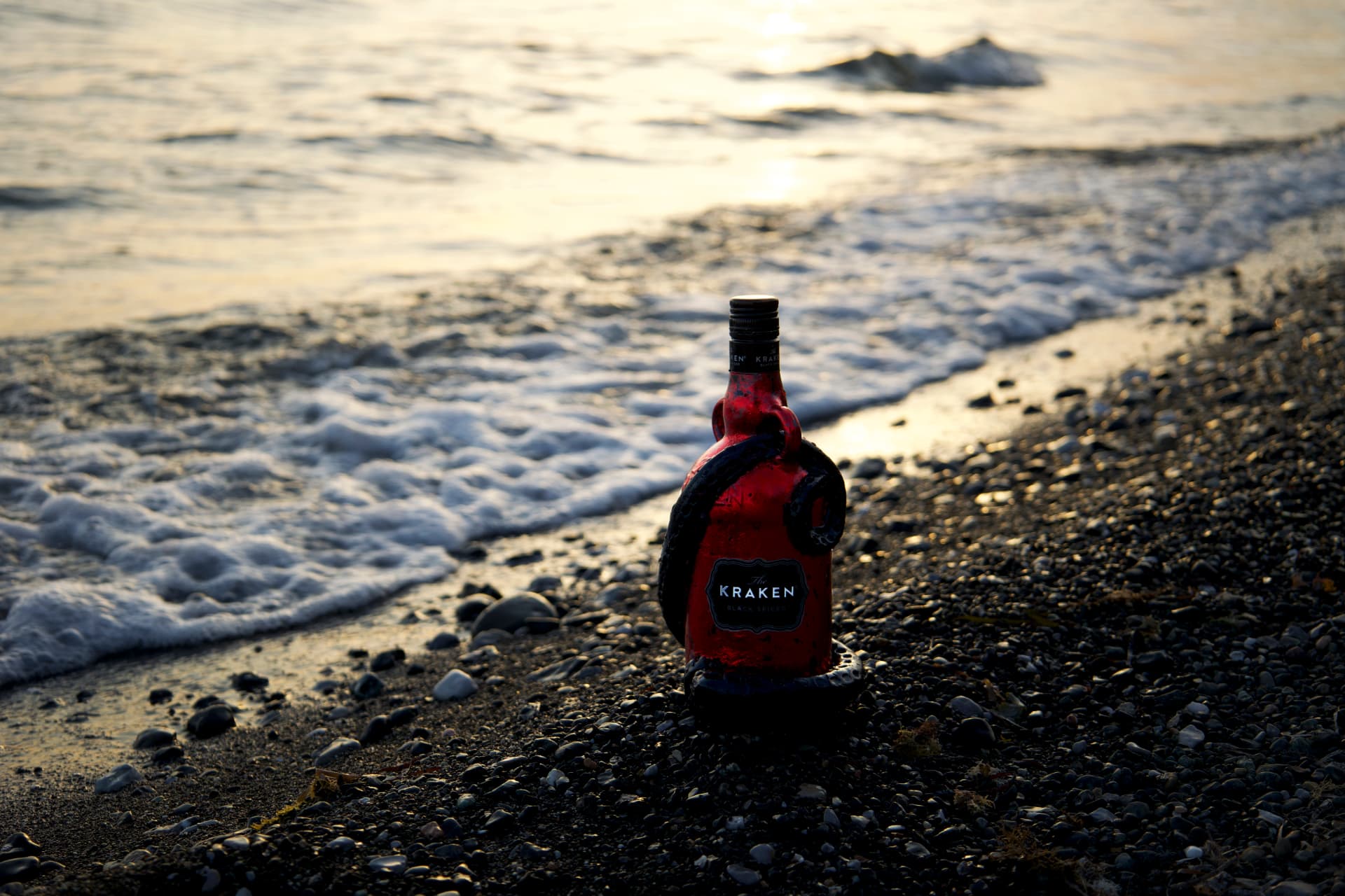 A red Kraken rum bottle standing on a pebble beach at sunset with gentle waves behind it.