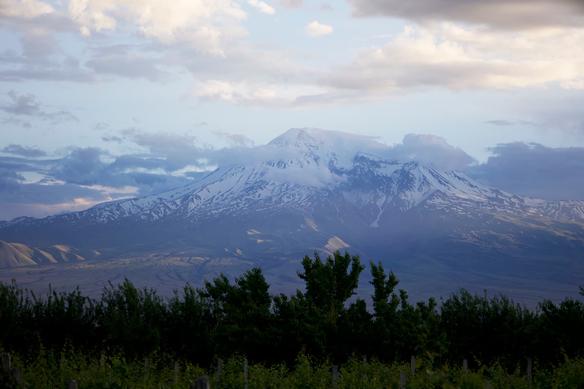 Snow-capped Mount Ararat rising above green hills and vineyards under a partly cloudy sky