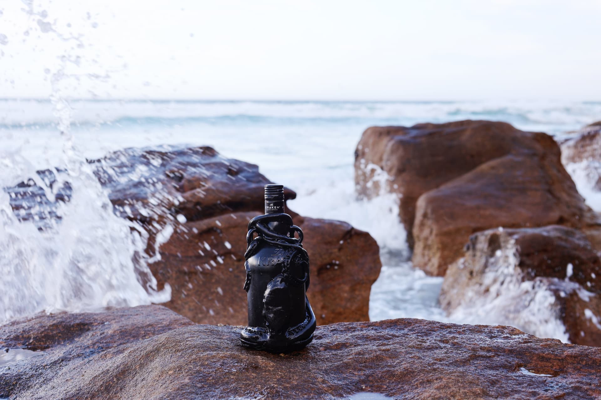 A black Kraken rum bottle sculpted with tentacles, standing on a rocky shore with waves crashing behind it.