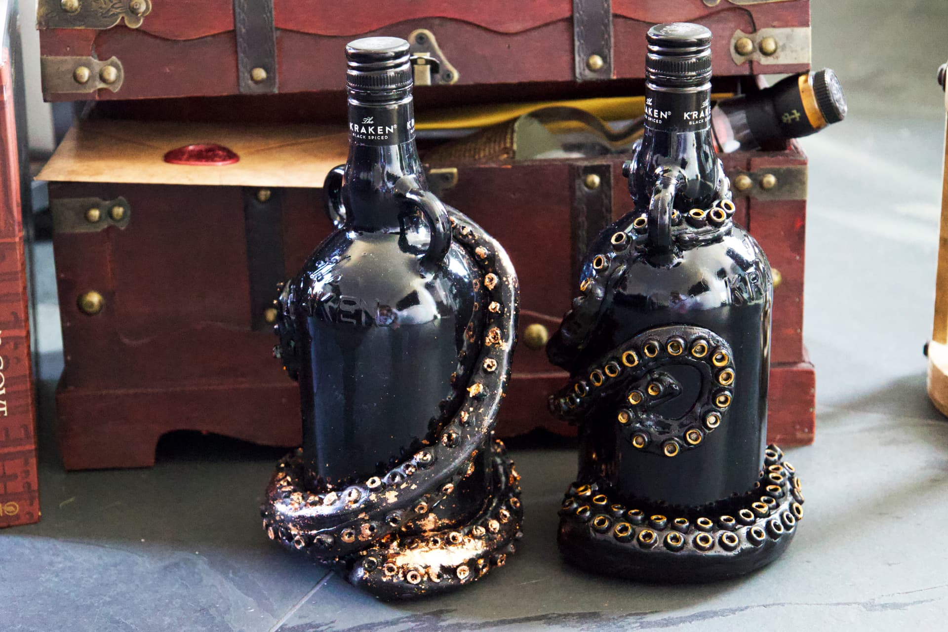 Two black Kraken rum bottles decorated with sculpted tentacles and gold-ringed suction cups, standing in front of vintage wooden chests.