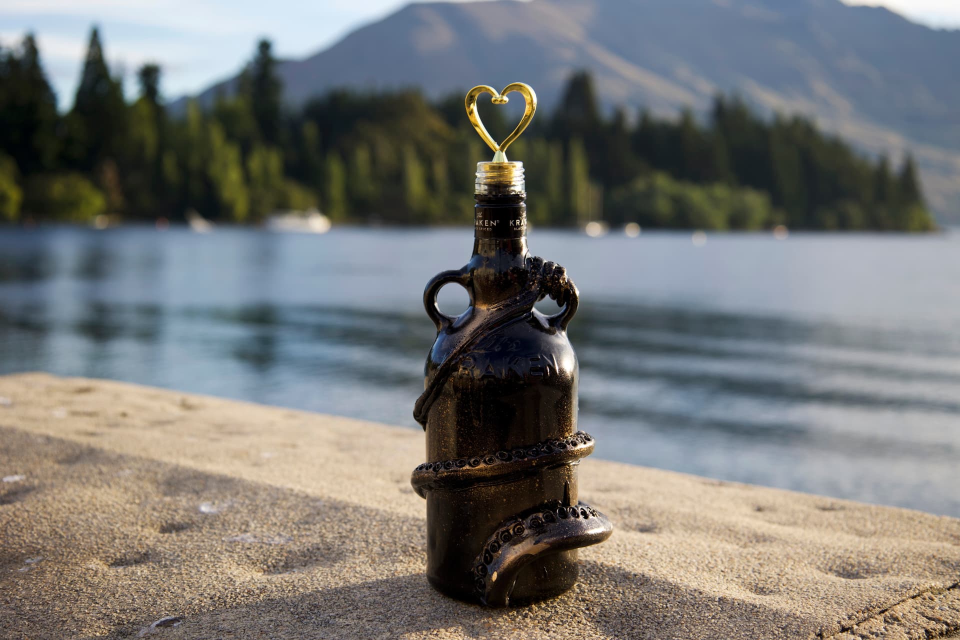 A black Kraken rum bottle decorated with sculpted tentacles and a golden heart-shaped wire, standing on a lakeside rock with mountains behind it.
