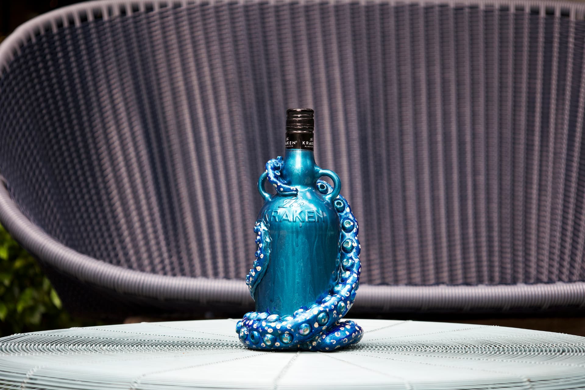 A teal Kraken rum bottle sculpted with bright blue tentacles and ringed suction cups, standing on a woven outdoor table.
