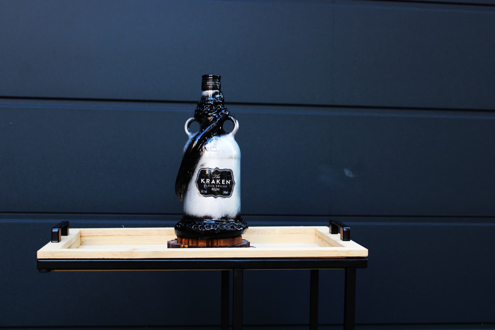 A silver-painted Kraken rum bottle with sculpted black tentacles, standing on a wooden tray against a dark background.